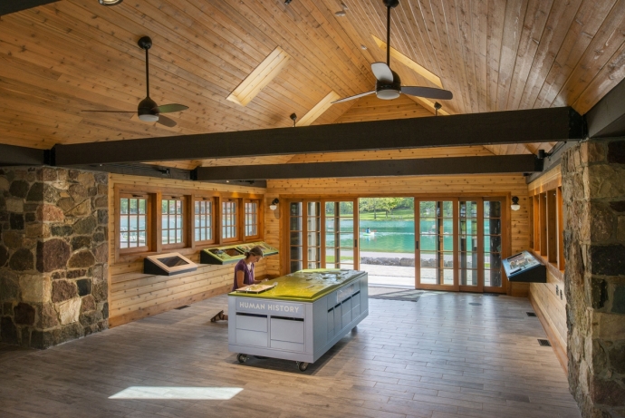 A woman looks at an interpretive display inside the environmental education center. The interior is clad with wood and stone and features large wood beams. The lake can be seen through the large doors at the back of the building.